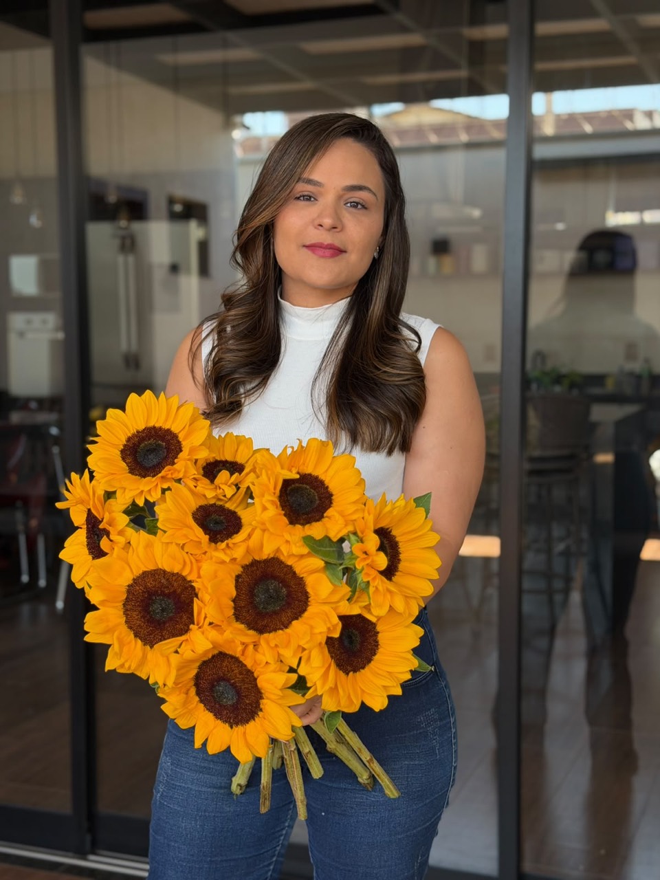 Priscilla Reis segurando um buquê de girassóis diante de portas de vidro.